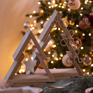 May include: Two wooden Christmas tree decorations. One is a simple triangle shape with a star ornament hanging from it. The other is a triangle with decorative gold rope accents. The background is a blurred Christmas tree with lights.