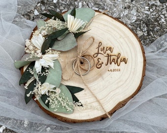 May include: A wooden ring bearer box with a natural wood finish. The box is decorated with dried eucalyptus leaves and white flowers. Two gold wedding bands are placed on the box. The box is engraved with the names "Luca & Italia" and the date "4.6.2022".