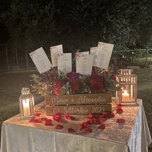 May include: A wooden box with the text "Marco e Alexandra 02 Settembre 2023" on it. The box is filled with red and purple flowers and has several white cards with text on them sticking out of the top.