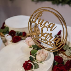 May include: A three-tiered wedding cake decorated with red and pink roses, white cotton balls, and a wooden sign that reads "Luca & Sara 21.12.2022".