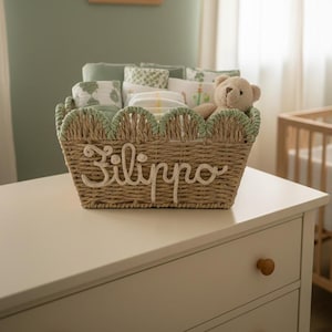 Può includere: Un cestino in vimini intrecciato con il nome "Filippo" in corsivo. Il cestino è pieno di articoli per bambini, tra cui pannolini e coperte. Un piccolo orsacchiotto è all'interno. Il cestino ha un bordo smerlato con finiture verdi.