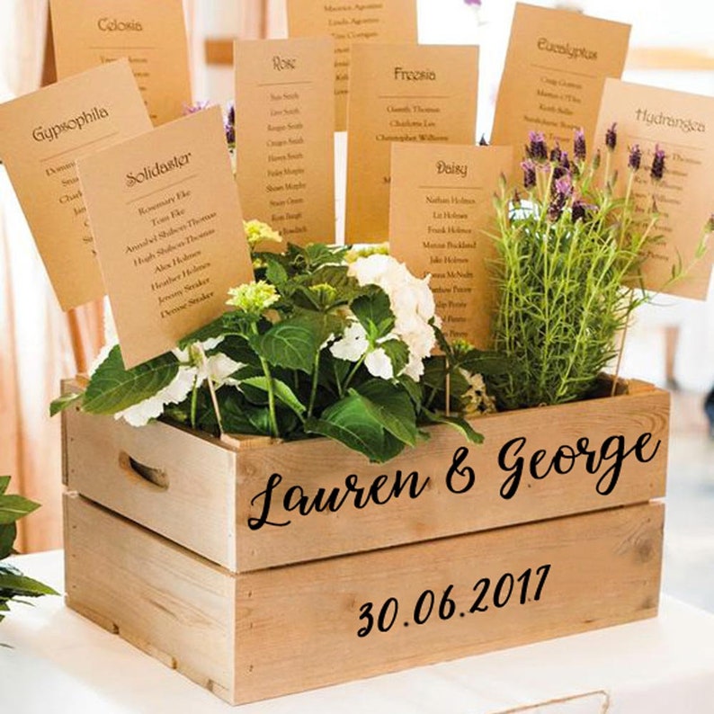 May include: A wooden crate with the names "Lauren & George" and the date "30.06.2017" written on it. The crate is filled with potted plants, including hydrangeas, lavender, and other greenery. The plants are labeled with brown paper cards with names like "Rose", "Freesia", and "Solidaster".