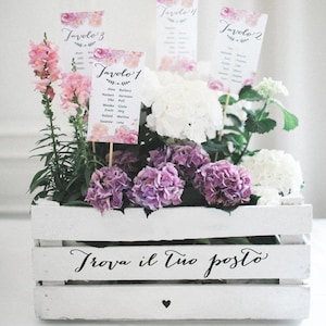May include: A white wooden crate with the text "Trova il tuo posto" in black script. The crate is filled with pink and white flowers and four cards with the text "Tavolo 1", "Tavolo 2", "Tavolo 3", and "Tavolo 4" in black script.