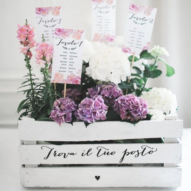 May include: A white wooden crate with the text "Trova il tuo posto" in black script. The crate is filled with pink and white flowers and four cards with the text "Tavolo 1", "Tavolo 2", "Tavolo 3", and "Tavolo 4" in black script.