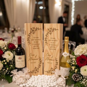 Puede incluir: Una mesa de recepción de bodas con una botella de vino tinto, una botella de champán y dos cajas de madera con diseños florales y texto en italiano. La mesa está decorada con flores blancas y rojas, y pequeñas bolsas de almendras confitadas.