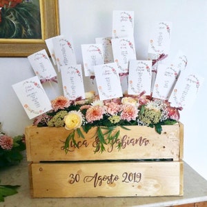 May include: A wooden crate with the names "Luca & Fabiana" and the date "30 Agosto 2019" written on it. The crate is filled with pink and white flowers and has small cards with floral designs and names written on them sticking out of the flowers.
