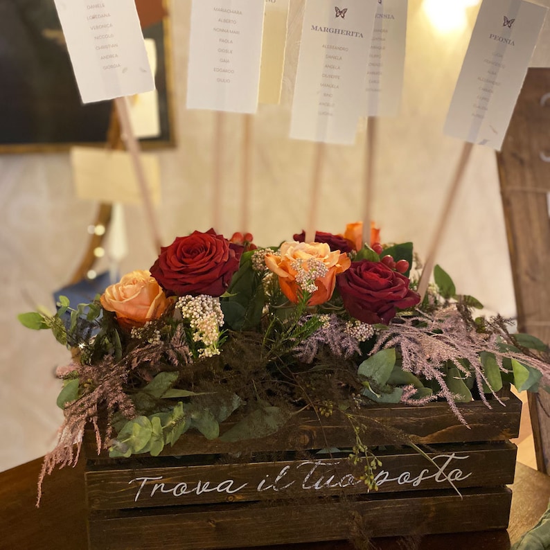 May include: A wooden crate with the text "Trova il tuo posto" written in white calligraphy. The crate is filled with a floral arrangement of red, orange, and pink roses, greenery, and sprigs of dried flowers.  The arrangement is topped with small white cards with names written on them.