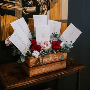 May include: A wooden crate with the names "Lica & Sara" and the date "22.12.2022" written on it. The crate is filled with red and pink roses and white cotton flowers. There are also several white cards with the word "Table" and a number on them.