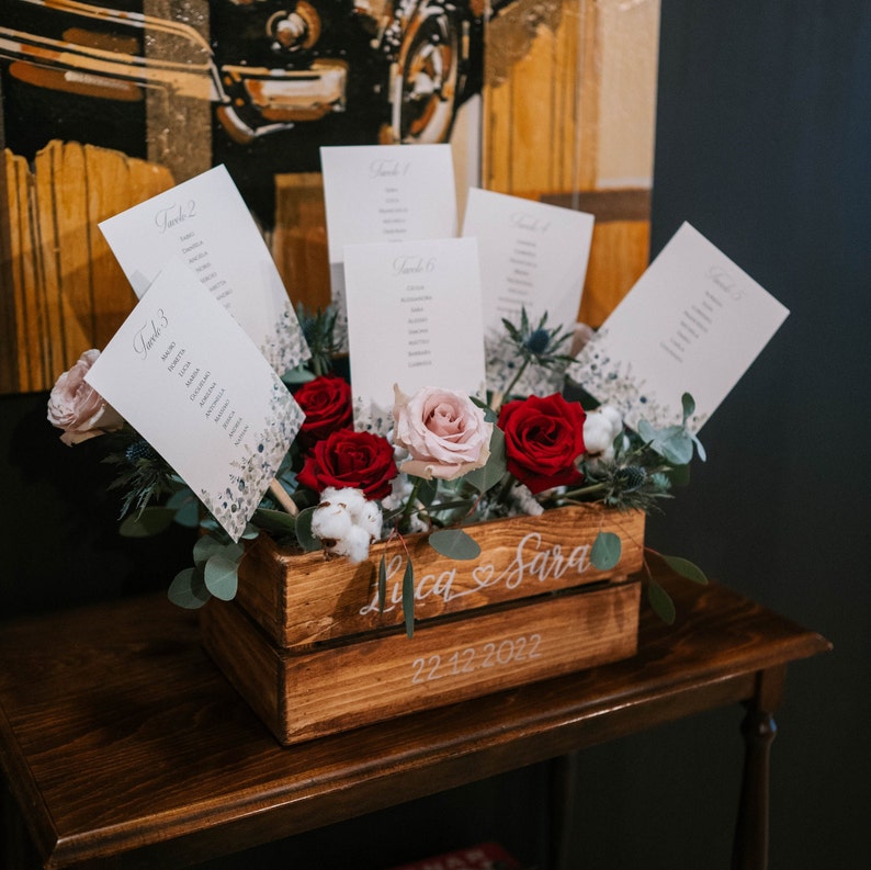 May include: A wooden crate with the names "Lica & Sara" and the date "22.12.2022" written on it. The crate is filled with red and pink roses and white cotton flowers. There are also several white cards with the word "Table" and a number on them.