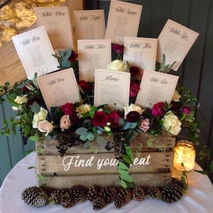 May include: A wooden crate filled with a floral arrangement of white, pink, and burgundy roses, greenery, and berries. The crate is decorated with the text "Find your eat" and has cards with table numbers and a menu card placed on top.