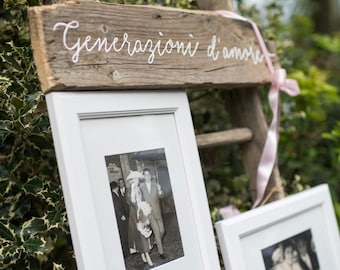 Può includere: Un cartello in legno rustico con il testo "Generazioni d'amore" in scrittura bianca. Il cartello è appeso a una scala di legno con due cornici bianche. Le cornici contengono fotografie in bianco e nero di una coppia.