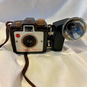 May include: A vintage Kodak Brownie Holiday Flash camera. The camera is brown and cream-colored with a flash attachment. The camera has a brown strap and the words "Brownie Holiday Flash Camera" are printed on the front.