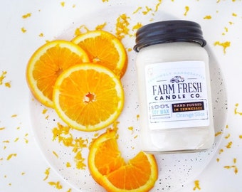 Peut inclure: Une bougie en pot blanche avec un couvercle noir, étiquetée "Farm Fresh Candle Co. Orange Slice", est posée sur une assiette blanche avec des tranches d'orange. La bougie est entourée de zeste d'orange. Fabriquée avec 100% de cire de soja et coulée à la main dans le Tennessee.