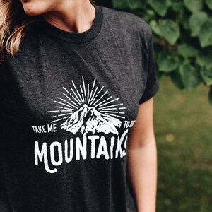 May include: A dark grey t-shirt with a white graphic design of a mountain range with a sunburst above it. The text "TAKE ME TO THE MOUNTAINS" is printed below the mountain graphic.