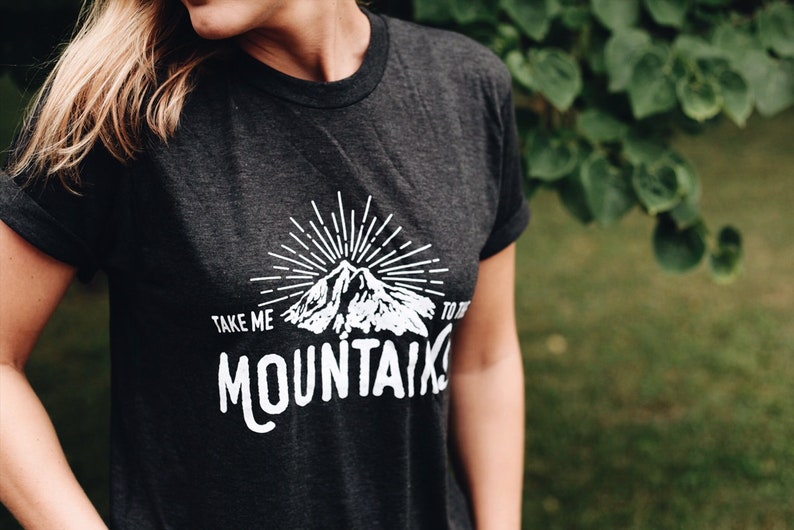 May include: A dark grey t-shirt with a white graphic design of a mountain range with a sunburst above it. The text "TAKE ME TO THE MOUNTAINS" is printed below the mountain graphic.