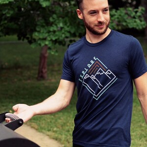 以下が含まれることがあります： 白いアウトラインで「EXPLORE」という文字と山々のグラフィックが描かれたネイビーブルーのTシャツ。山々は茶色、青、緑の色合いです。