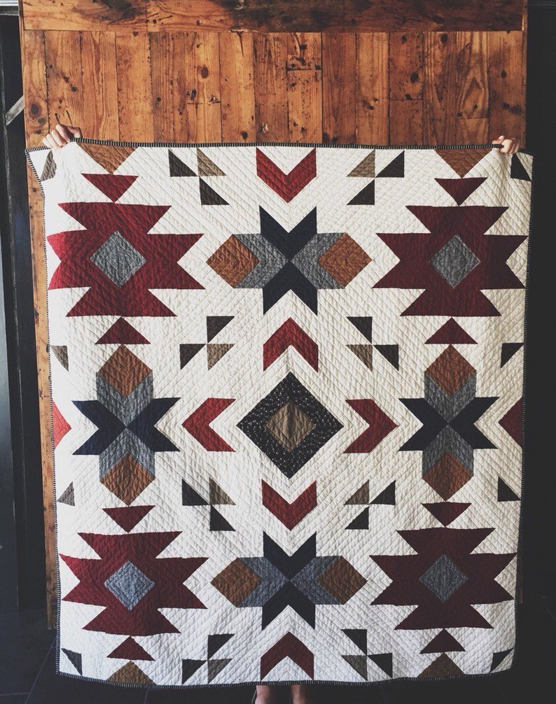 Pu&ograve; includere: Un quilt bianco con un motivo geometrico in tonalit&agrave; di rosso, marrone, nero e grigio. Il quilt presenta un design a stella ripetuto e forme a freccia.