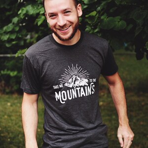 May include: A dark grey t-shirt with a white graphic design of a mountain with sun rays and the text "Take me to the mountains".