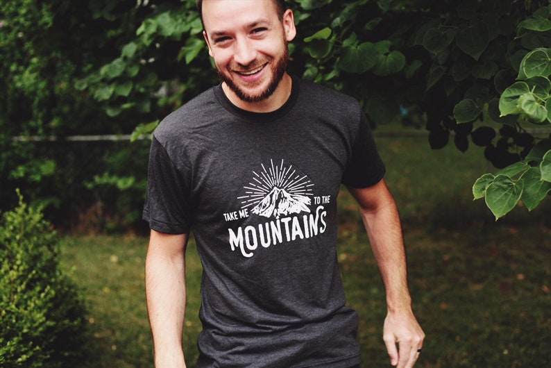 May include: A dark grey t-shirt with a white graphic design of a mountain with sun rays and the text "Take me to the mountains".