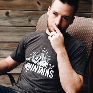 May include: A man wearing a dark grey t-shirt with a white graphic that says "Take me to the mountains" and a mountain silhouette. He is sitting in a chair and holding a bottle of beer.