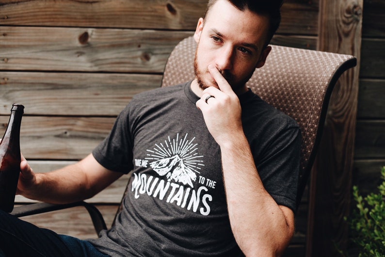 May include: A man wearing a dark grey t-shirt with a white graphic that says "Take me to the mountains" and a mountain silhouette. He is sitting in a chair and holding a bottle of beer.