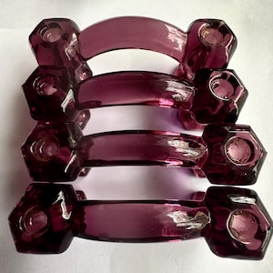 Op de afbeelding: Vier antieke, amethistkleurige glazen servetringen. Elke ring heeft een gebogen handvat dat twee zeshoekige uiteinden verbindt. Het glas is doorschijnend, waardoor licht erdoorheen kan schijnen, en het algehele ontwerp is elegant en vintage.