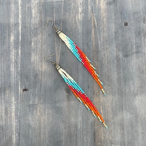 May include: A pair of long, beaded earrings with a white, turquoise, orange, and gold color scheme. The earrings are made with small, round beads and have a simple, elegant design.