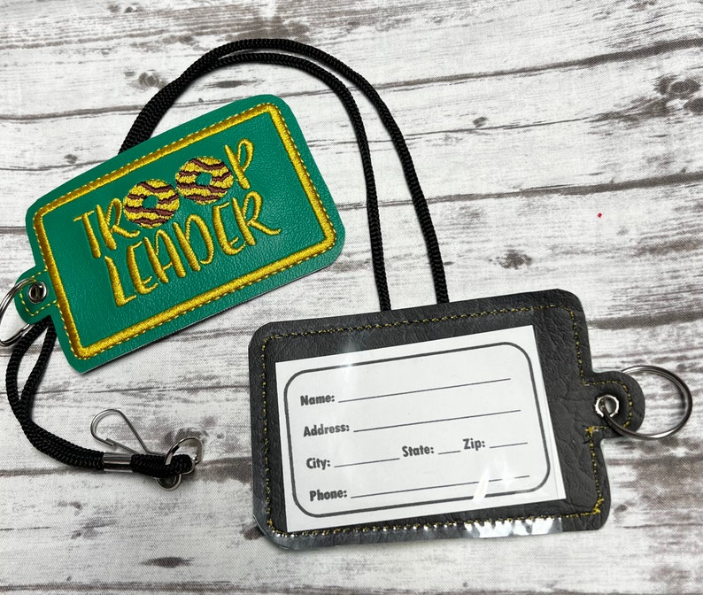 May include: Green and gold embroidered badge with the text "Troop Leader" and two brown and gold cookie images. A black lanyard with a black and gold ID holder with a clear plastic window for a name tag.