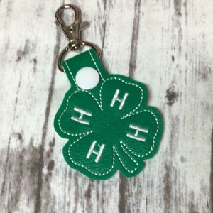 May include: Green leather keychain shaped like a four-leaf clover with white stitching and the letters "H" embroidered on each leaf.