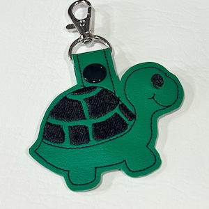 May include: Green turtle-shaped keychain with a black patterned shell design. The keychain has a silver-colored clasp and a black button detail. The turtle has a smiling face.