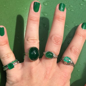 May include: A hand wearing four rings with green gemstones. The rings are set in silver and have a delicate design. The hand is also wearing green nail polish.