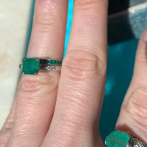 May include: Two emerald rings on a finger. The top ring features a square emerald stone with smaller emerald stones set in the band. The bottom ring features a round emerald stone with smaller diamonds set in the band.