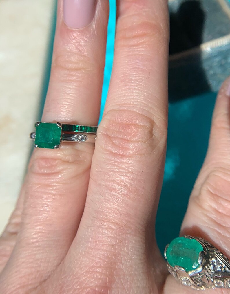 May include: Two emerald rings on a finger. The top ring features a square emerald stone with smaller emerald stones set in the band. The bottom ring features a round emerald stone with smaller diamonds set in the band.