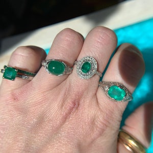May include: Three silver rings with emerald gemstones. The rings are on a person's hand. The largest ring has a round emerald surrounded by smaller diamonds. The other two rings have square and oval emeralds.