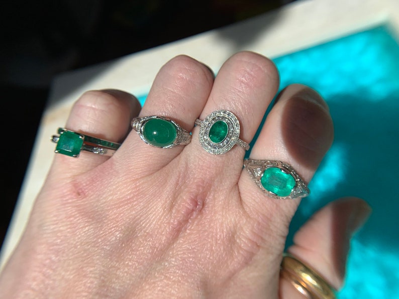 May include: Three silver rings with emerald gemstones. The rings are on a person's hand. The largest ring has a round emerald surrounded by smaller diamonds. The other two rings have square and oval emeralds.
