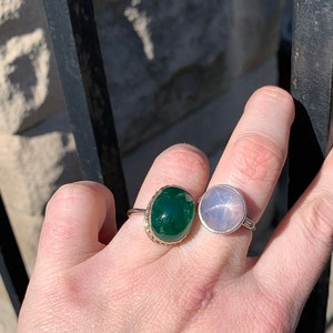 May include: Two rings with oval-shaped gemstones. The ring on the left has a dark green stone set in a gold band. The ring on the right has a light pink stone set in a silver band.