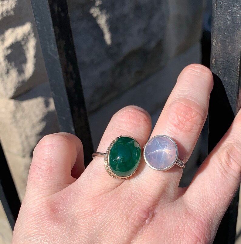 May include: Two rings with oval-shaped gemstones. The ring on the left has a dark green stone set in a gold band. The ring on the right has a light pink stone set in a silver band.