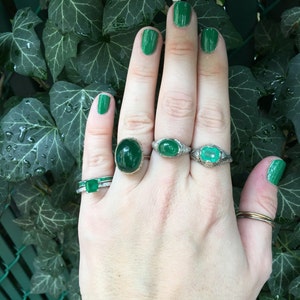 May include: A hand with green nail polish is wearing four rings with emerald green gemstones. The rings are set in silver and have a delicate design.