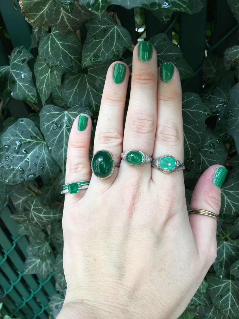May include: A hand with green nail polish is wearing four rings with emerald green gemstones. The rings are set in silver and have a delicate design.