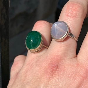 May include: Two rings with oval-shaped gemstones. The ring on the left has a dark green gemstone with a gold setting. The ring on the right has a light blue gemstone with a silver setting.