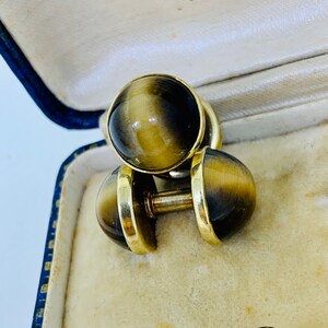 K&ouml;nnte beinhalten: Ein Paar goldene Manschettenkn&ouml;pfe mit Tigeraugen-Steinen. Die Manschettenkn&ouml;pfe befinden sich in einer blau-wei&szlig;en Schachtel mit dem Text "6, Bridge Street, Inverness".