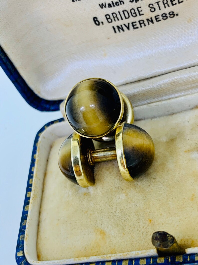 K&ouml;nnte beinhalten: Ein Paar goldene Manschettenkn&ouml;pfe mit Tigeraugen-Steinen. Die Manschettenkn&ouml;pfe befinden sich in einer blau-wei&szlig;en Schachtel mit dem Text "6, Bridge Street, Inverness".