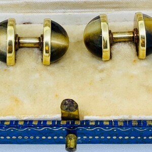 K&ouml;nnte beinhalten: Ein Paar goldene Manschettenkn&ouml;pfe mit braunen und goldenen Tigeraugen-Steinen. Die Manschettenkn&ouml;pfe befinden sich in einer blauen und goldenen Schachtel.