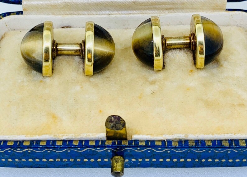 K&ouml;nnte beinhalten: Ein Paar goldene Manschettenkn&ouml;pfe mit braunen und goldenen Tigeraugen-Steinen. Die Manschettenkn&ouml;pfe befinden sich in einer blauen und goldenen Schachtel.