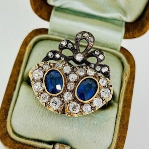 May include: An antique gold-tone brooch with two oval, deep blue gemstones surrounded by small, clear gemstones. A black bow design tops the brooch, which is displayed in a vintage jewelry box.