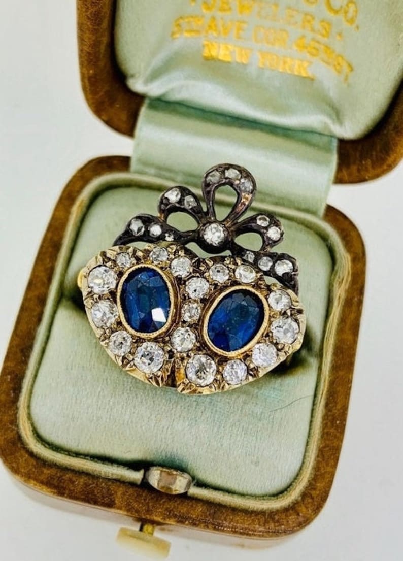 May include: An antique gold-tone brooch with two oval, deep blue gemstones surrounded by small, clear gemstones. A black bow design tops the brooch, which is displayed in a vintage jewelry box.