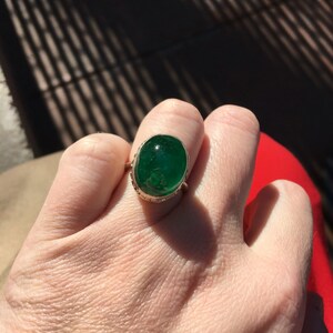 May include: A gold ring with a large oval emerald green gemstone set in a bezel setting. The ring is on a person's finger.
