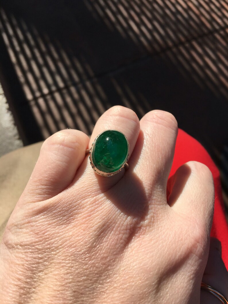 May include: A gold ring with a large oval emerald green gemstone set in a bezel setting. The ring is on a person's finger.