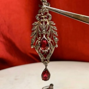 Może przedstawiać: Para antycznych srebrnych kolczyków z czerwonymi kamieniami szlachetnymi. Kolczyki mają delikatny wzór filigranowy z małymi błyszczącymi kamieniami.