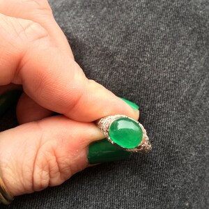 May include: A silver ring with a green oval gemstone set in a bezel setting. The ring is surrounded by small, clear gemstones.